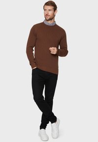 Brun ribbad tröja med rund halsringning, kombinerad med svarta slim-fit byxor och vita sneakers. En anmärkningsvärd detalj är en logotypdetalj.