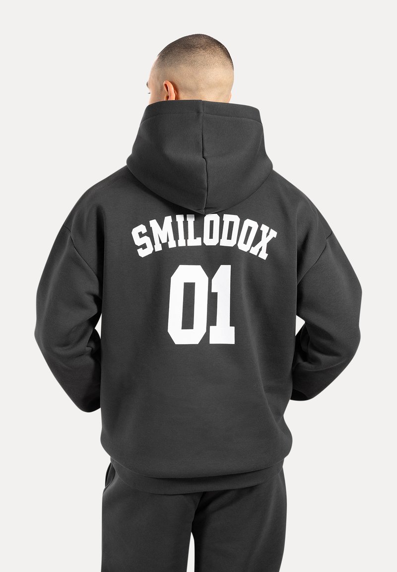 Szara bluza z kapturem z dużą białą grafiką na plecach, z napisem "SMILODOX 01". Posiada pojemny kaptur i ściągacze na końcach rękawów.