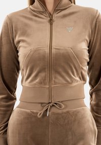 Giacca zip-up in velluto marrone con vestibilità strutturata, vita elastica, tasche frontali e un piccolo logo triangolare decorativo sul petto.