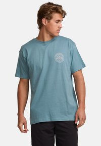 Light blue cotton t-shirt with short sleeves, crew neck, and a round logo printed on the chest featuring mountains and water design.