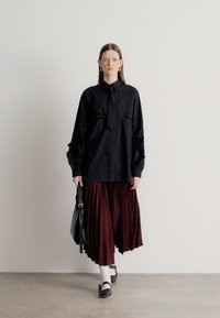 Črna srajca s tipi gumbi in dvema žepoma na prsih, kombinirana s bordo rdečo plisirano krilo. Model drži črno torbico, nosi bele nogavice in temne čevlje.