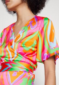Femme portant une robe portefeuille à manches courtes avec des tourbillons abstraits vibrants en orange, rose, vert, violet et bleu clair sur fond blanc.