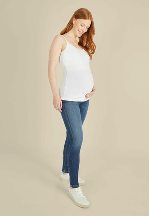Gravid kvinde med rødt hår iført en hvid tanktop, blå jeans og hvide sneakers, der blidt holder om sin mave og smiler.
