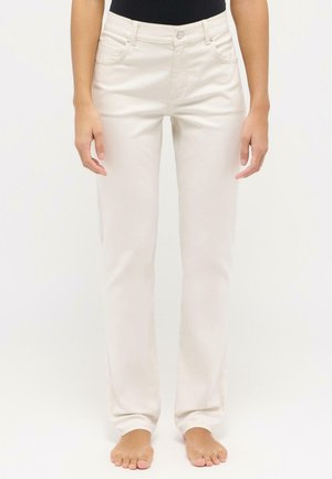 Kobieta ubrana w kremowe dżinsy o prostych nogawkach i czarną bluzkę, stojąca boso na jednolitym białym tle.