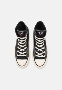 Zapatillas altas negras con costuras blancas, ojales dorados y cordones blancos. Tiene acentos de bordado colorido y una puntera de goma.