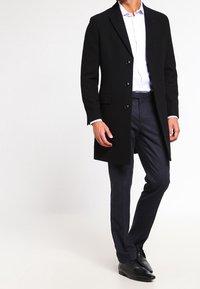 Man in een zwarte knopenjas, witte overhemd, donkergrijze broek en zwarte nette schoenen, met één been iets naar voren.