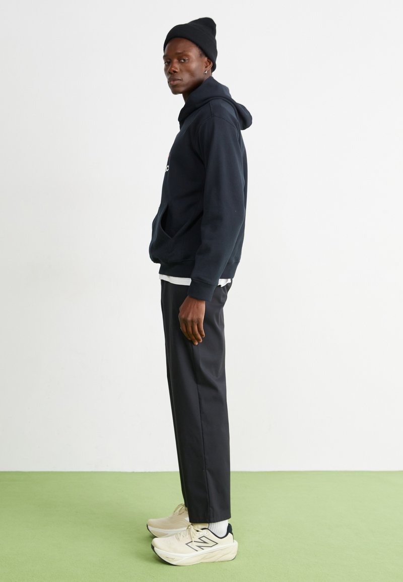Schwarzer Hoodie, darunter ein weißes Hemd, schwarze Hose, beige Sneakers. Das Model trägt eine schwarze Mütze und steht auf einem grünen Boden.
