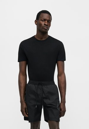 Man with dark skin wearing a fitted black short-sleeve t-shirt and black drawstring shorts standing against a plain white background.