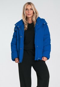 Niebieska kurtka ocieplana zapinana na zamek błyskawiczny i napy, z kapturem, kieszeniami bocznymi oraz pikowanym wzorem. Nosić ją na czarnej bluzce.