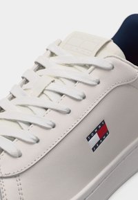 Λευκό δερμάτινο αθλητικό παπούτσι με λευκά κορδόνια και μικρό λογότυπο Tommy Jeans σε κόκκινο, λευκό και ναυτικό μπλε στη πλάγια επιφάνεια.