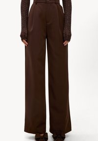 Un pantalon large marron présente une taille haute, des plis et un tissu lisse et texturé. Il est complété par un haut ajusté avec des manches transparentes à pois.