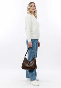 Brown leather handbag with a smooth texture, curved shape, and silver hardware. Model wears a white sweater and light blue jeans.