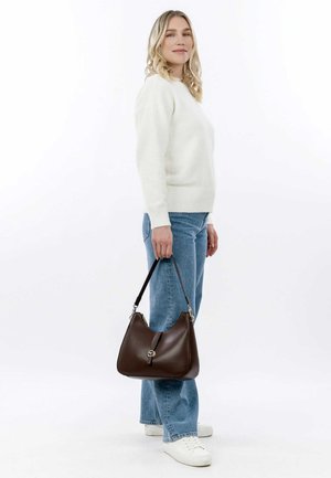 Brown leather handbag with a smooth texture, curved shape, and silver hardware. Model wears a white sweater and light blue jeans.