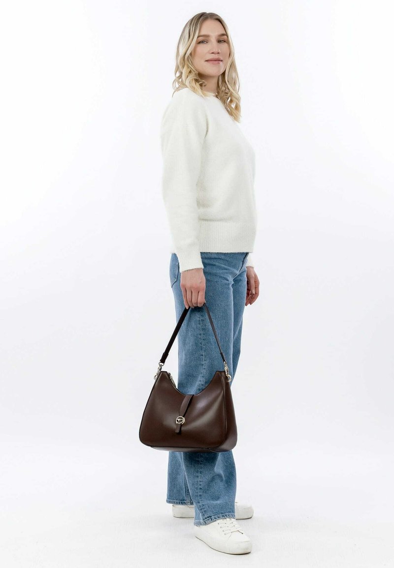 Brown leather handbag with a smooth texture, curved shape, and silver hardware. Model wears a white sweater and light blue jeans.