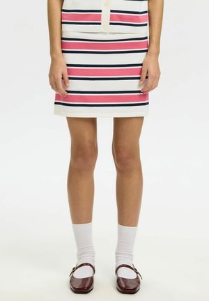 Nainen, joka pukeutunut valkoiseen, vaaleanpunaiseen ja tummansiniseen raidalliseen hameeseen, valkoiset sukat ja tummanpunaiset Mary Jane -kengät, seisoo kädet rentoina sivuilla.