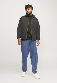Fekete cipzáras dzseki, zöld pulóver alatta, laza kék farmernadrág és fehér sneaker. A modell egy sima háttér előtt áll, enyhén oldalra fordulva.