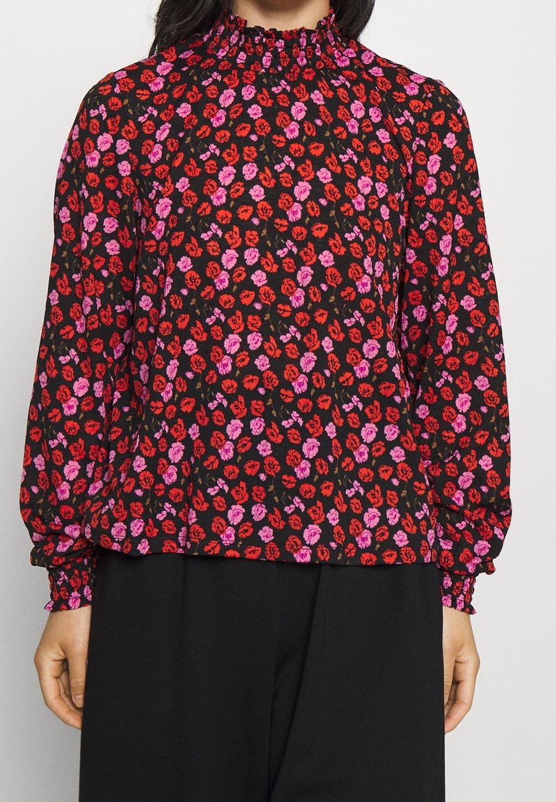 Blouse noire à manches longues avec un col smocké haut, ornée d'un motif floral dans des teintes de rouge et de rose, avec des poignets froncés et une coupe décontractée.