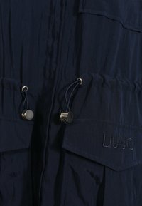 Giacca leggera blu navy con finitura testurizzata, dotata di due tasche frontali, dettagli con coulisse e accenti metallici. Brand: LIU JO.