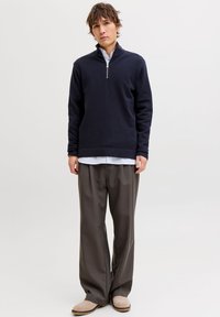 Πλεκτή μπλούζα σε ναυτικό μπλε με φερμουάρ και μακριά μανίκια, κατασκευασμένη από μαλακό ύφασμα, συνδυασμένη με φαρδιά γκρι παντελόνια και ανοιχτό μπεζ παπούτσια, με μινιμαλιστικό σχεδιασμό.