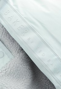Mjukt grått fleece-tyg med en texturerad yta, som har en ljusblå, gummibelagd etikett präglad med "BAKER by TED BAKER."