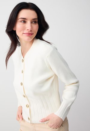 Woman with dark hair wearing a white buttoned cardigan and beige pants, posing with hands in pockets, looking to the side.