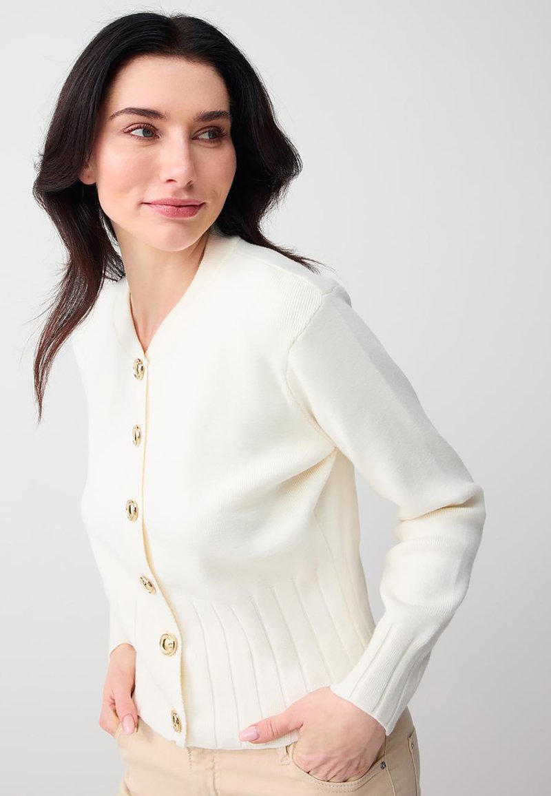Femme aux cheveux foncés portant un cardigan blanc boutonné et un pantalon beige, posant les mains dans les poches et regardant sur le côté.