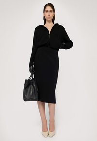 Svart zip-front tröja med huva, svart figurnära pennkjol, svart läderhinkväska och nakna spetsiga klackar. Släta texturer genomgående.