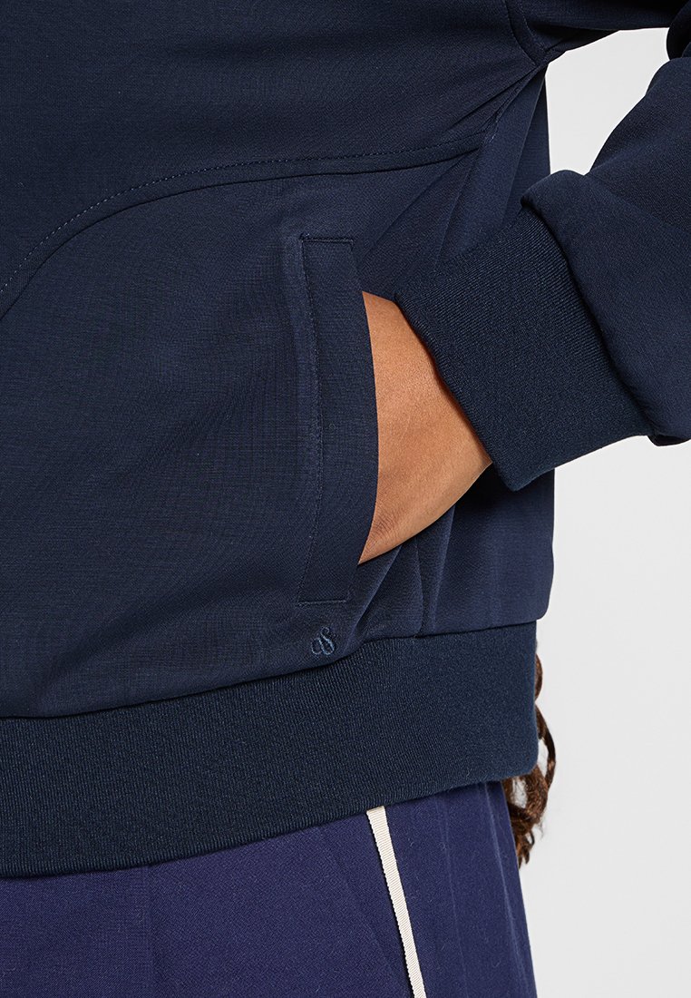 Main dans la poche d'une veste bleu marine avec des poignets et un ourlet côtelés, associée à un pantalon sombre avec une rayure verticale blanche.