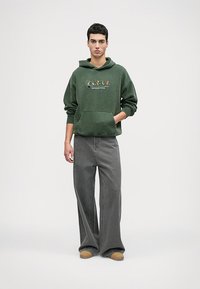 Zaļa flīsa kapuce ar priekšējo kabatu un grafisku apdruku, apvienota ar brīviem pelēkiem denima biksēm un brūnām kurpēm. Minimalistisks dizains.