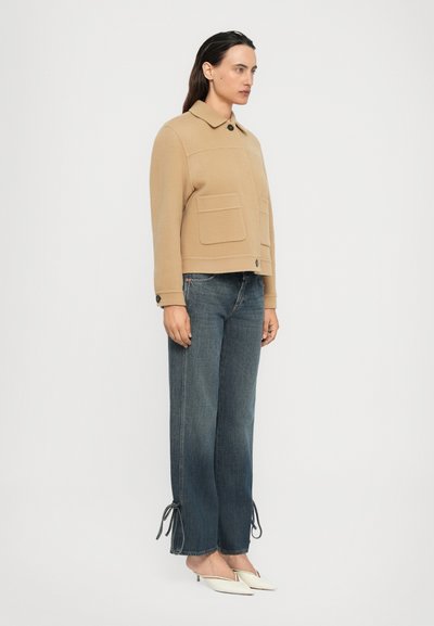 Frau in einer beigen, knopfgeknöpften Jacke mit großen Vorderaschen, dunkelblauen, weit geschnittenen Jeans mit Bändern an den Säumen und weißen, hochhackigen Mules.