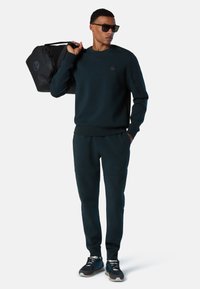 Sudadera verde oscura y joggers de un material suave, combinados con zapatillas deportivas negras y gafas de sol negras. Lleva un bolso de deporte negro.