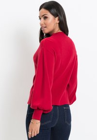 Maglione rosso lavorato a maglia con collo rotondo, maniche a palloncino e design cropped, abbinato a jeans blu scuro. Tessuto morbido e stile aderente.