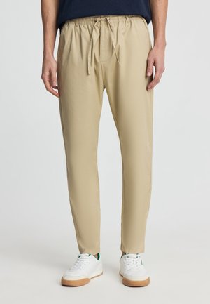 Uomo che indossa pantaloni beige con coulisse e sneakers bianche, in piedi davanti a uno sfondo chiaro e uniforme.