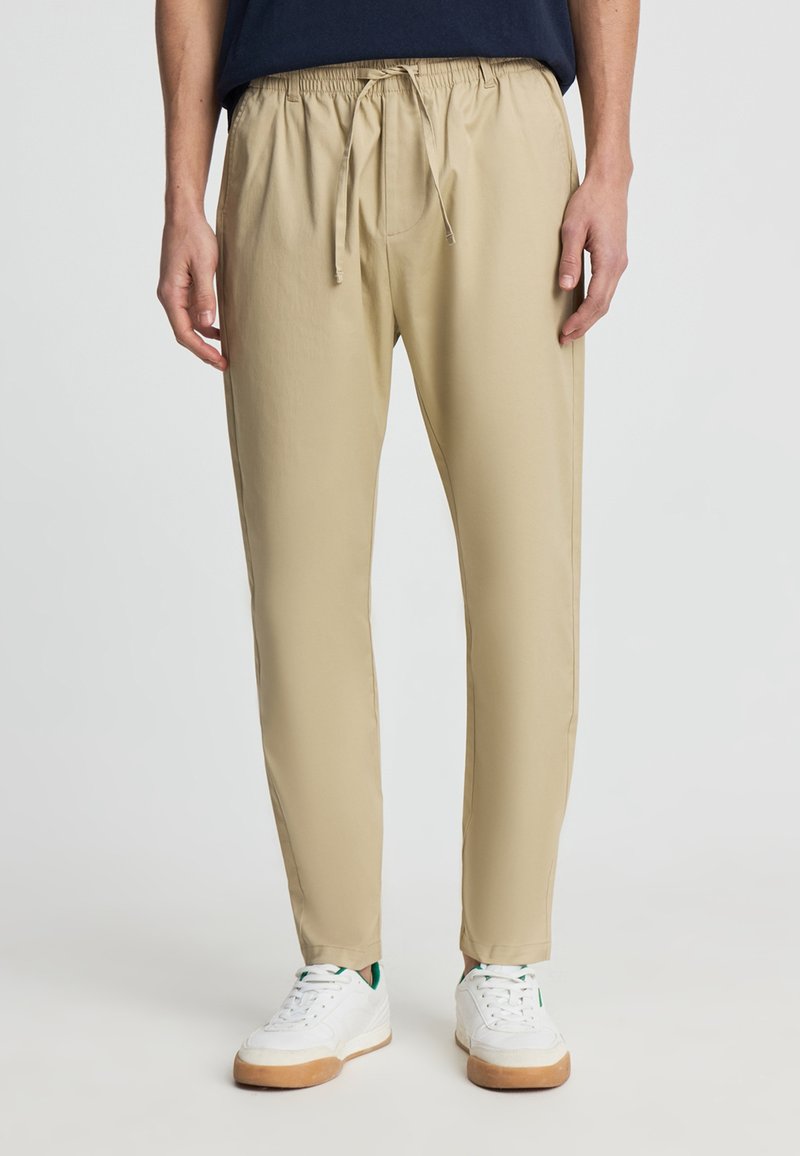 Uomo che indossa pantaloni beige con coulisse e sneakers bianche, in piedi davanti a uno sfondo chiaro e uniforme.
