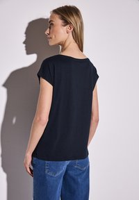 Černý top s krátkým rukávem a volným střihem, s kulatým výstřihem, kombinovaný s světle modrými džíny. Materiál je hladký a mírně texturovaný.