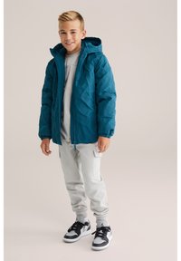 Chaqueta acolchada en color verde azulado con un patrón texturizado, cierre frontal y capucha. Combinada con pantalones de chándal gris claro y zapatillas en blanco y negro.