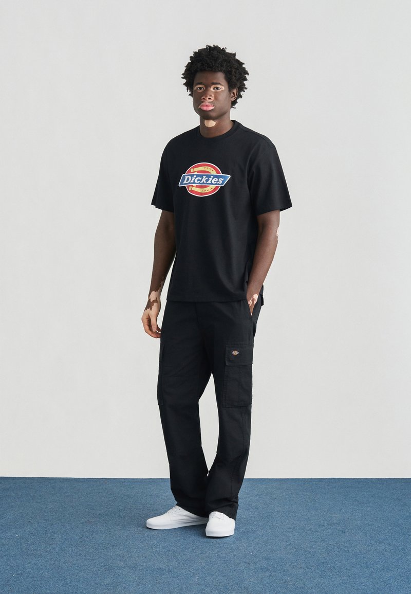 T-shirt nera di cotone con logo "Dickies" in un design circolare colorato. Abbinate a pantaloni cargo neri e scarpe da ginnastica bianche. Sfondo semplice.