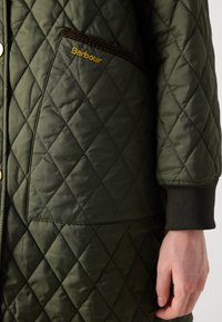 Gequiltte groene jas met een ruitpatroon, bruine koordafwerking op de zak en een gouden "Barbour" logo op de linkerkant van de borst.