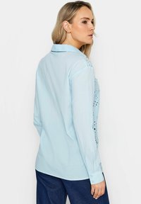 Camicia azzurro chiaro a maniche lunghe con un delicato motivo di pizzo sulle spalle, dotata di colletto classico e polsini con bottoni. Tessuto liscio e leggero.