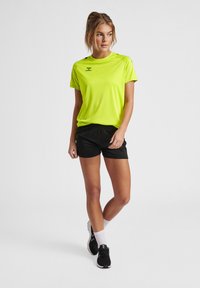 Neon grøn kortærmet atletisk skjorte med logo, parret med sorte shorts. Modellen har sorte sneakers og hvide sokker på.