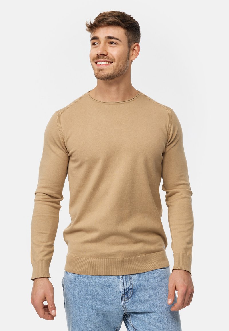 Beige strikket sweater med rund hals, ribbede manchetter og talje. Texturen er glat med en tætsiddende design. Parret med lyseblå jeans.