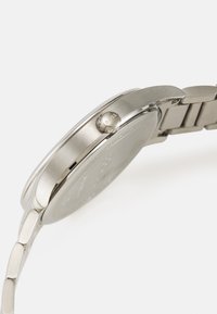 Reloj de acero inoxidable plateado con una caja redonda, un acabado cepillado suave y una corona texturizada que muestra el nombre de la marca.