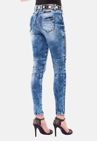 Jeans de mezclilla azul ajustados con áreas desgastadas, acentos laterales con cremallera y un cinturón negro con tachuelas. Los zapatos de tacón alto tienen adornos de picos.