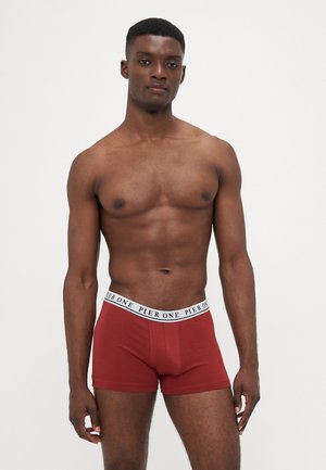 Sportlicher, dunkelhäutiger Mann steht vor einem neutralen Hintergrund und trägt rote Boxershorts mit einem weißen Bund, auf dem "Pier One" steht.