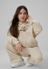 Jeune fille aux longs cheveux bruns portant un sweat à capuche beige et un pantalon, accroupie tenant des écouteurs blancs sur un fond gris uni.