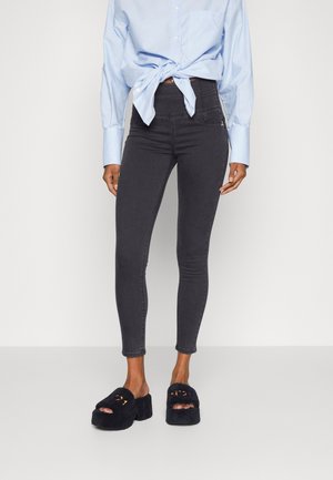 Femme portant un jean skinny noir taille haute, une chemise bleu clair à manches longues nouée à la taille, et des sandales à plateforme noires et duveteuses.