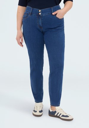 Person iført blå højtaljede jeans med frontknapper og lommer, kombineret med hvide og marineblå sneakers, stående på en lys baggrund.