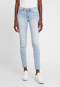 Jeans ajustados de color azul claro en tela de mezclilla con un patrón desvanecido, diseño de cinco bolsillos y pasadores para cinturón. Combinados con zapatillas blancas.