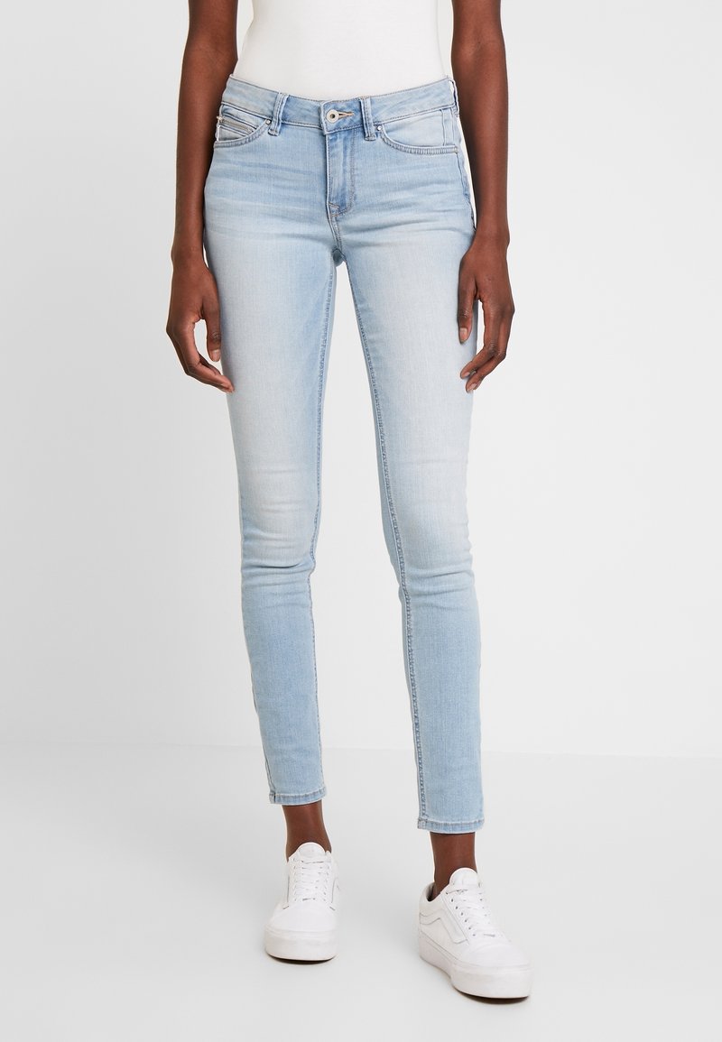 Jeans ajustados de color azul claro en tela de mezclilla con un patrón desvanecido, diseño de cinco bolsillos y pasadores para cinturón. Combinados con zapatillas blancas.