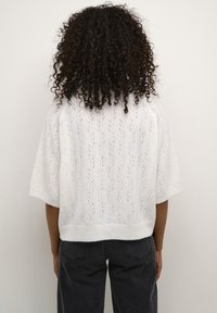 Pull en tricot blanc avec un motif de feuilles, à manches courtes et coupe décontractée, porté avec un pantalon gris foncé, vu de dos.